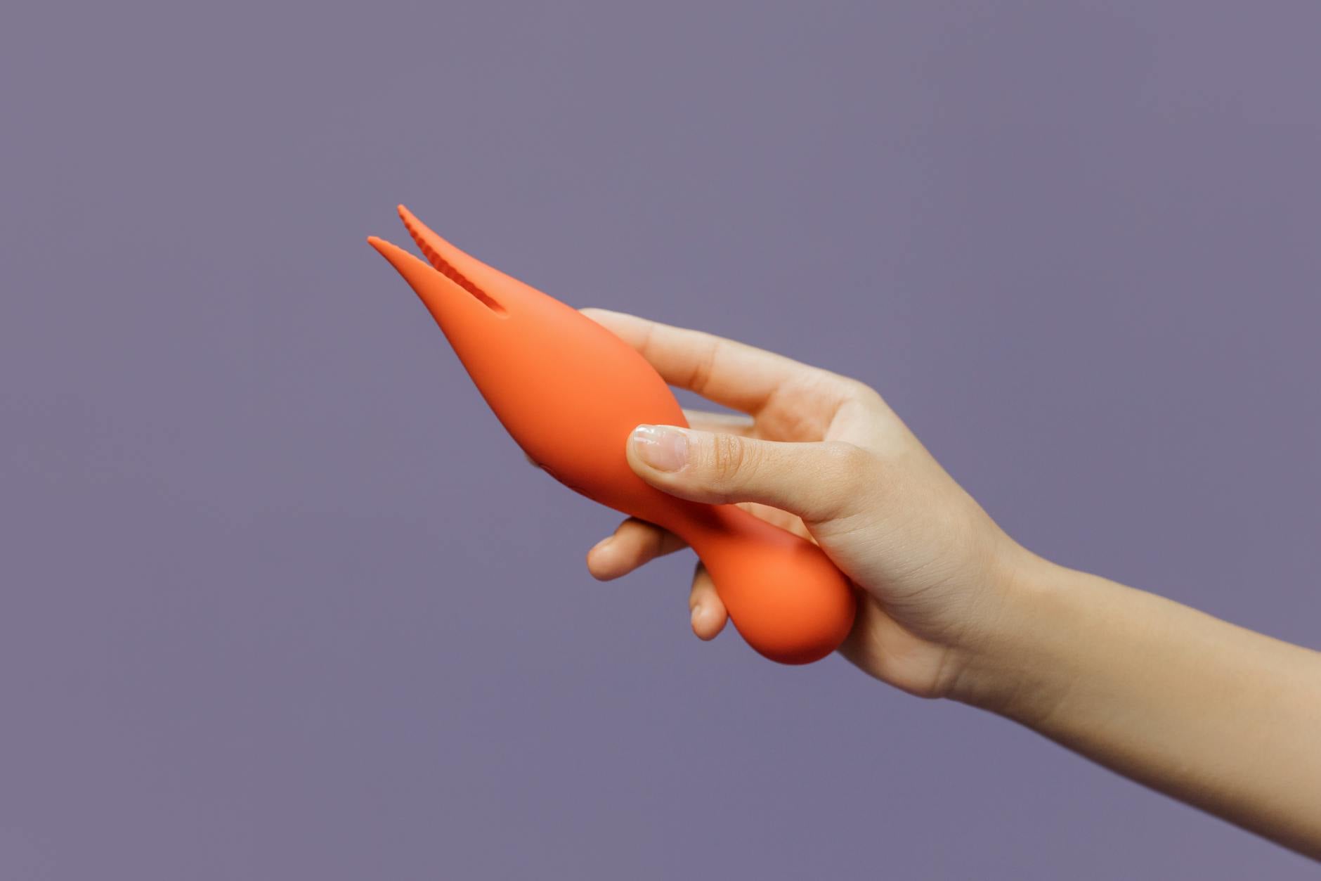 Close-up of a hand holding an orange vibrator against a minimalistic purple backdrop