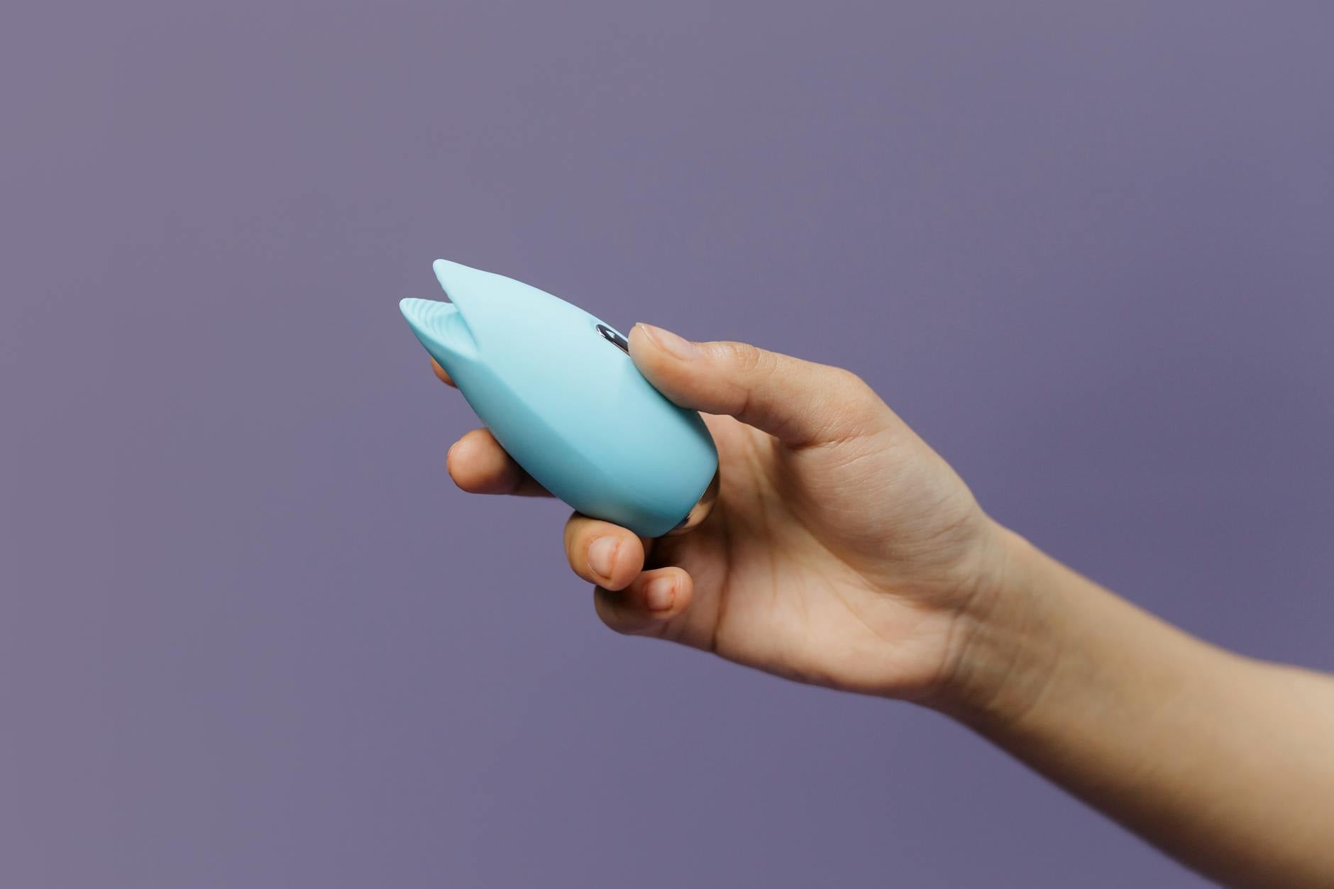 A blue silicone clitoral vibrator held gently in hand against a purple background