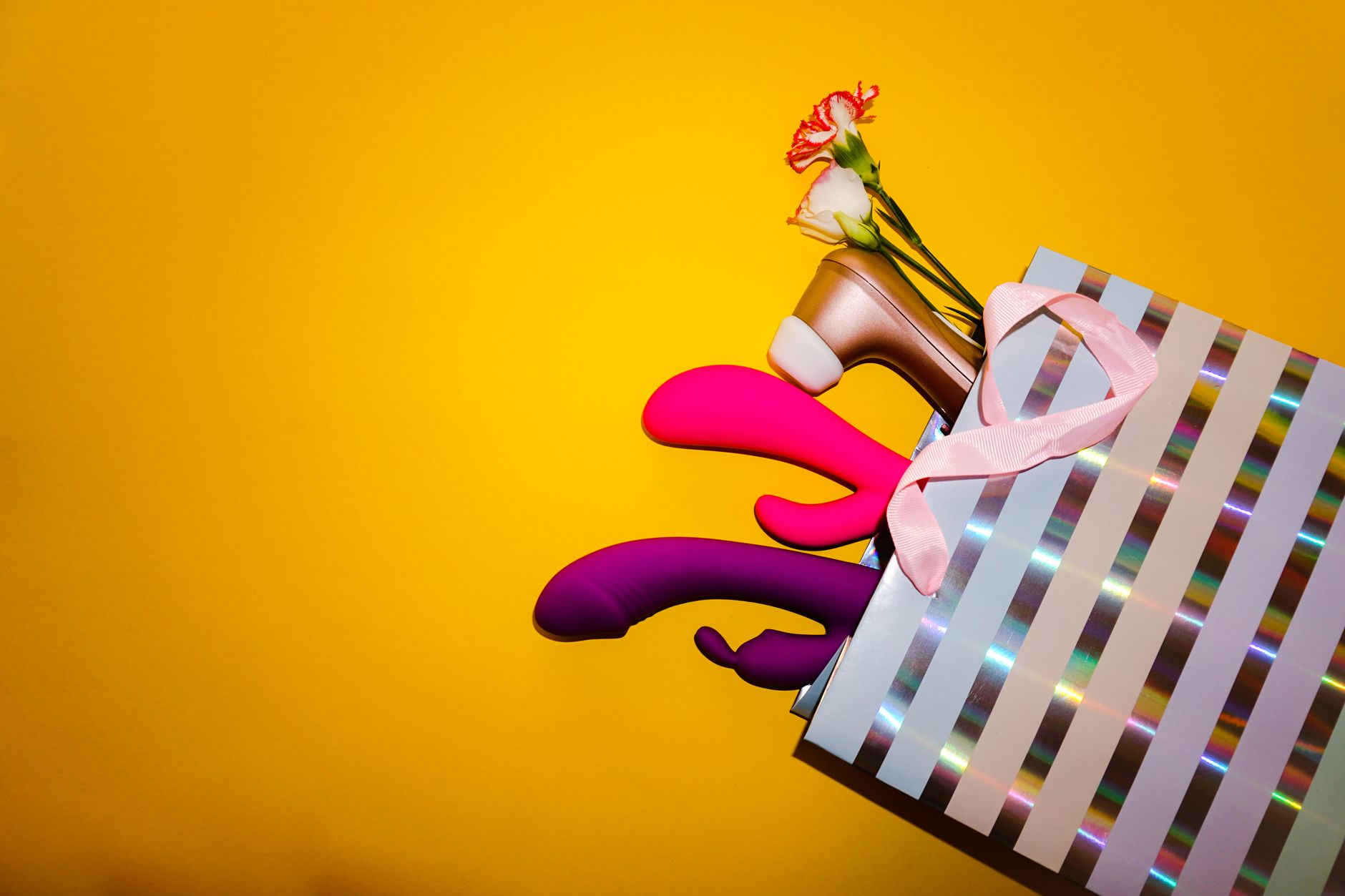 Colorful vibrators with flowers displayed in a holographic gift bag against a bold yellow background.